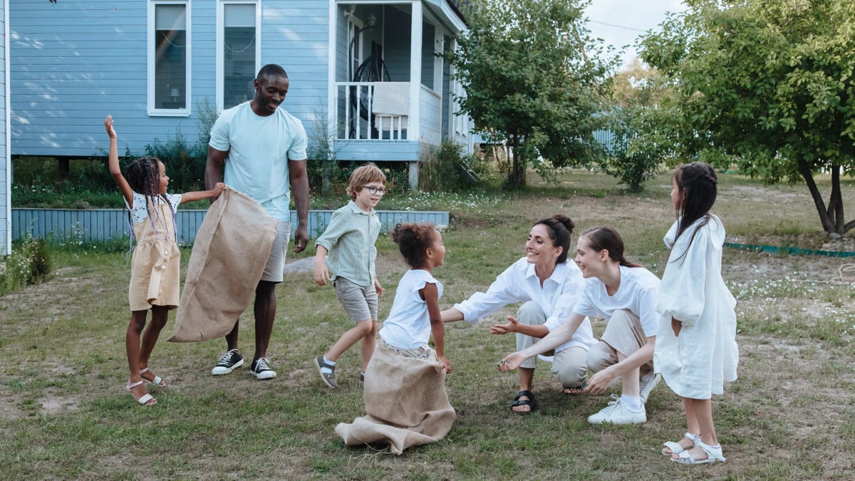 Children and adults enjoying a sack race beside a blue house on a sunny day.